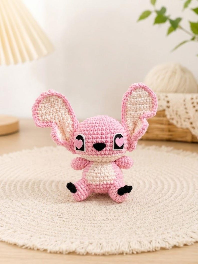 May include: A handmade crochet plush toy in the shape of a pink and white cartoon character with large ears. The toy has black eyes with heart-shaped pupils and black feet. It is sitting on a cream-colored, round rug.
