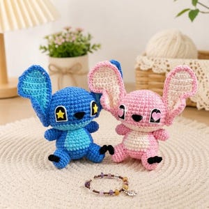 May include: Two crocheted plush toys, one blue and one pink, resembling a cartoon character. The blue toy has a yellow star on its ear, while the pink one has a heart. A beaded bracelet with a charm is in front of them. The toys are on a white, textured surface.