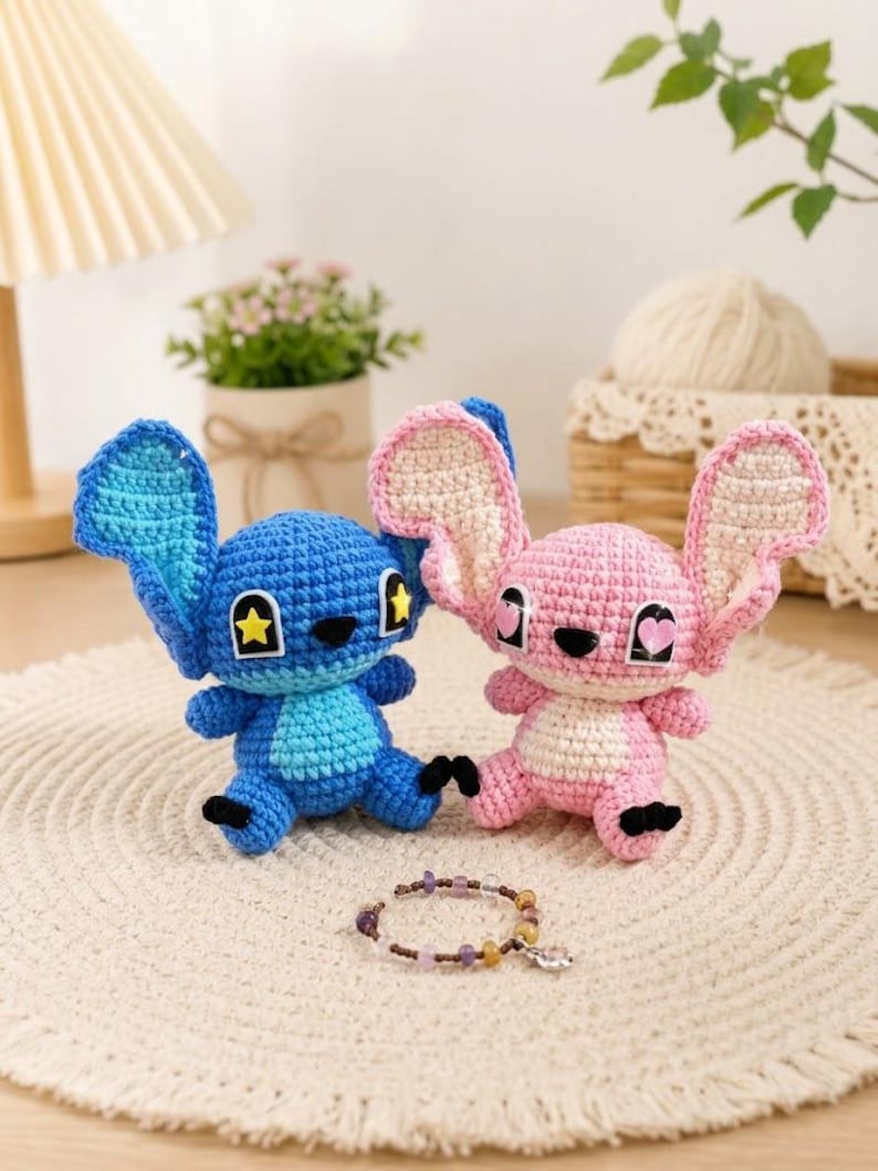 May include: Two crocheted plush toys, one blue and one pink, resembling a cartoon character. The blue toy has a yellow star on its ear, while the pink one has a heart. A beaded bracelet with a charm is in front of them. The toys are on a white, textured surface.