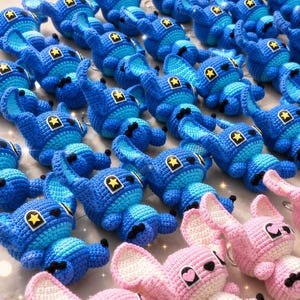 May include: Crocheted blue and pink character plushies with black accents and star-shaped details. The blue plushies have a yellow star on their heads, while the pink ones have heart-shaped details. The plushies are arranged in rows.