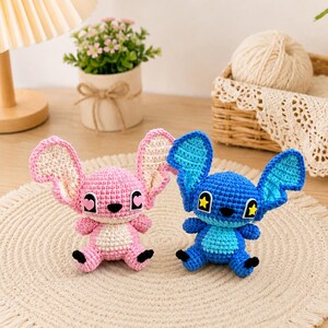 May include: Two crocheted plush toys, one pink and one blue, resembling a cartoon character. The pink toy has light pink ears and a white belly, while the blue toy has blue ears and yellow star accents. Both have black eyes and feet.