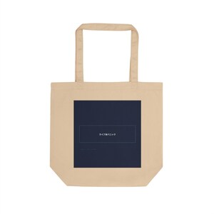 May include: A beige tote bag featuring a dark blue square with Japanese text. The bag has two beige handles and is made of a durable fabric. The design is simple and modern, suitable for carrying items. The bag is a neutral colour.