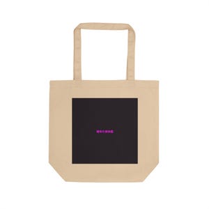 May include: Beige tote bag with a black square in the centre. The black square has pink Japanese text. The bag has two beige handles and is made of a canvas-like material. The bag is empty.