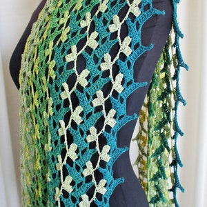 May include: A green and yellow crocheted shawl with a leafy pattern. The shawl is draped over a black mannequin.