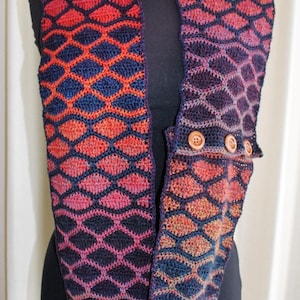 May include: A crocheted infinity scarf with a geometric pattern in shades of red, orange, purple, and blue. The scarf has two wooden buttons on the side.