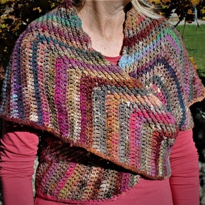 Puede incluir: Un chal de ganchillo multicolor con un patrón a rayas en tonos de marrón, rosa, morado y azul. El chal está drapeado sobre los hombros de una persona y se usa como una envoltura.