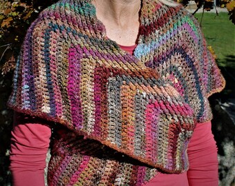 Può includere: Un scialle all'uncinetto multicolore con un motivo a righe in tonalità di marrone, rosa, viola e blu. Lo scialle è drappeggiato sulle spalle di una persona e viene indossato come un avvolgimento.