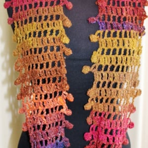 May include: A handmade crochet scarf in a spectrum of colors, including red, orange, yellow, and purple. The openwork design features a scalloped edge, creating a textured and visually appealing accessory. The scarf is displayed on a black mannequin.