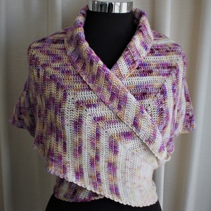 Puede incluir: Un chal de ganchillo en tonos de morado, blanco y amarillo. El chal está drapeado sobre un maniquí y tiene un diseño de cuello en V.