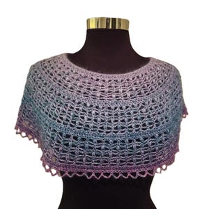 May include: A crochet poncho featuring a gradient color pattern that transitions from light purple at the neckline to blue and deep violet at the hem. The garment is crafted with an openwork stitch pattern and finished with a scalloped edge.
