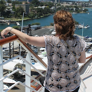 Puede incluir: Una mujer lleva una blusa de ganchillo con un patrón de espiral en beige y blanco. La blusa es sin mangas y tiene un corte holgado. La mujer está de pie en una cubierta con vistas a un puerto con un puente en el fondo.