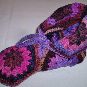 以下が含まれることがあります： 紫、ピンク、茶色の色合いのクロシェスリッパ。スリッパはさまざまなステッチで作られており、ユニークな質感のデザインです。