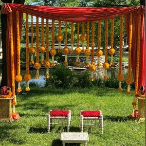 May include: An outdoor wedding setup with a red and orange draped backdrop adorned with strings of orange and white flowers. Two small red and white patterned stools and a small table sit on the green grass.