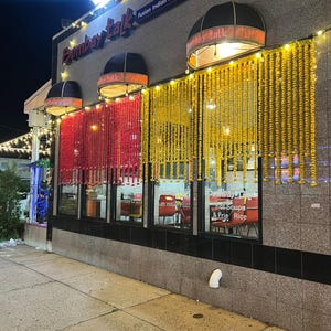 May include: Exterior view of the Bombay Talk restaurant at night. The building has a gray facade with red and yellow floral garlands hanging in front of the windows. The restaurant's name is displayed in a stylized font.
