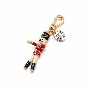 May include: A gold-coloured metal keyring featuring a miniature toy soldier. The soldier wears a red coat, black hat, and white trousers. A small gold tag reads "LIMITED EDITION 2006".