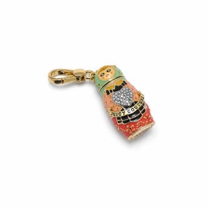May include: A gold-tone metal charm shaped like a nesting doll with enamel details in mint green, peach, and red. The doll features a crystal-encrusted heart, black accents, and a banner with the text Juicy Couture attached to a lobster clasp.