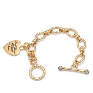 Può includere: Bracciale dorato con design a catena e chiusura a leva. Un ciondolo a forma di cuore con la scritta "Royal Couture Juicy" pende dalla catena. Il bracciale ha una finitura lucida.