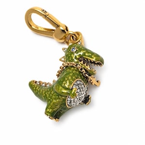 Pode incluir: Um pingente de dinossauro em metal dourado com esmalte verde, barriga incrustada com cristais brancos, um pequeno olho de cristal azul e uma corrente dourada fina com um pingente de chave, preso a um fecho lagosta dourado.