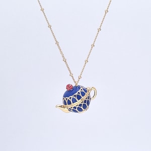 May include: A gold-toned necklace featuring a pendant shaped like a blue teapot with gold filigree and a red gem on top. The teapot has a small handle and spout. The chain is a delicate gold color.