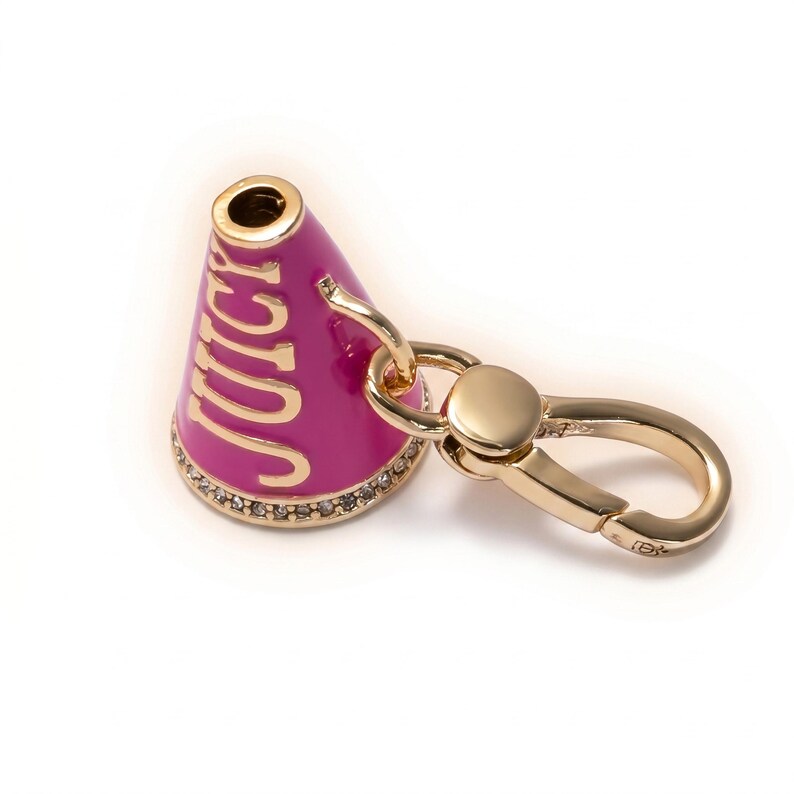 May include: A gold-toned metal charm in the shape of a cheerleader megaphone. The megaphone is magenta with the word "JUICY" in gold lettering. The base is encrusted with small, clear crystals, and it has a gold clasp.
