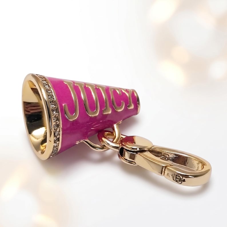 May include: A gold-colored keychain with a pink megaphone charm. The megaphone has the word "JUICY" in gold lettering. The megaphone is accented with small, clear crystals around the rim. The keychain has a gold-colored clasp.