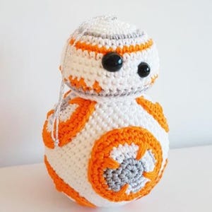 Peut inclure: Ornement fait main en crochet représentant BB-8. La sphère blanche est ornée de détails orange et gris, avec des yeux noirs en forme de boutons et une ficelle argentée pour suspendre. Décoration sur le thème de Star Wars.