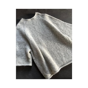 Puede incluir: Un suéter de punto gris claro con cuello redondo y mangas cortas. El suéter está hecho de un hilo suave y texturizado y tiene un ajuste relajado. La prenda se muestra sobre una superficie oscura.