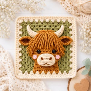 May include: A square crochet granny square featuring a three-dimensional Highland cow head in the center. The cow has golden-brown yarn hair, cream-colored horns, black eyes, and pink cheeks. The background is olive green with a cream border.