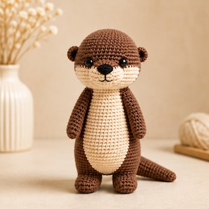 Puede incluir: Un peluche de nutria tejido a ganchillo a mano con un cuerpo marrón oscuro y un vientre y cara de color crema en contraste. La figura tiene orejas pequeñas y redondas, ojos negros de seguridad, una nariz negra bordada y una cola larga.