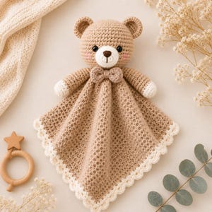 May include: A light brown crochet security blanket featuring a teddy bear head with black eyes, a dark brown nose, and pink cheeks. The bear wears a small bow tie, and the blanket has a white scalloped edge with white tips on the bear's paws.