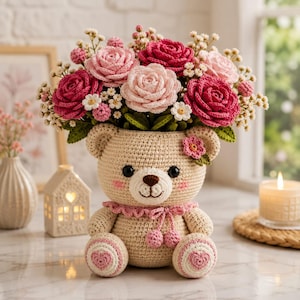 May include: A beige crocheted teddy bear planter holding a bouquet of pink and dark pink crocheted roses with small white filler flowers. The bear features a pink ruffled collar with two small pom-poms, pink heart-shaped foot pads, and a small flower.