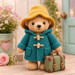 Puede incluir: Un oso de peluche tejido a ganchillo con un sombrero amarillo y una trenca de color verde azulado con dos botones de palanca claros. El oso tiene ojos negros y nariz bordada, y sostiene una pequeña maleta rectangular verde con correas marrones.