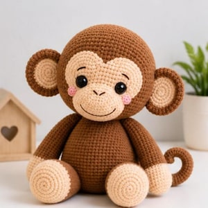 Op de afbeelding: Een gehaakte apenknuffel in bruin en beige met grote ronde oren, zwarte veiligheidsoogjes, een geborduurde glimlach en kleine roze cirkelvormige accenten op de wangen. De aap heeft een gekrulde staart en lichtgekleurde handen en voeten.