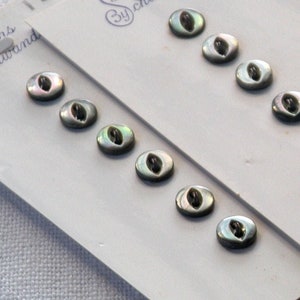 May include: A pack of 10 iridescent mother-of-pearl buttons with a silver finish. The buttons are oval shaped and have a small hole in the center. The buttons are labeled "Buttons by Schwanda".