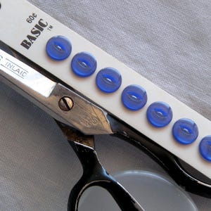 May include: A close-up of a package of seven blue, oval-shaped buttons, each 11mm (7/16") in size, with a pair of black-handled scissors. The package is labeled "BASIC".