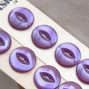 May include: Close-up of a set of iridescent purple buttons. Each button is round with a central, eye-shaped opening for thread. The buttons have a pearlescent sheen, reflecting light. The buttons are arranged on a cream-colored card.