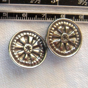 May include: Two silver-colored, round buttons with a wheel-like design. Each button has a central starburst pattern surrounded by radiating lines and a beaded outer edge. The buttons are next to a ruler.