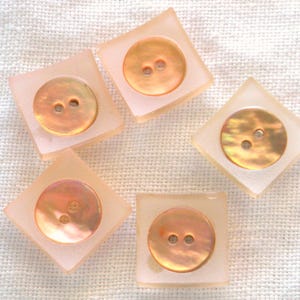 May include: Five square, light peach-colored buttons with a round, iridescent, two-hole center. The buttons are arranged on a textured, off-white fabric. The buttons are a soft, muted color.