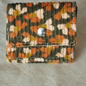 Op de afbeelding: Een klein rechthoekig corduroy tasje met een herhalend bloemenpatroon in tinten olijfgroen, gebrand oranje en crème. Het tasje wordt gesloten met een enkele, centraal geplaatste zilverkleurige metalen drukknoop en heeft zichtbare witte stiksels.