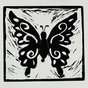 Puede incluir: Una ilustración en grabado de bloque en blanco y negro de una mariposa centrada dentro de un borde cuadrado. La mariposa presenta alas simétricas con patrones de remolinos y formas negras sólidas sobre un fondo blanco texturizado y tallado a mano.