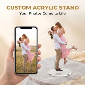 May include: A clear acrylic stand with a photo of a couple kissing, and a smartphone displaying the same image. The stand has a circular base and the text "CUSTOM ACRYLIC STAND" and "Your Photos Come to Life" in gold.