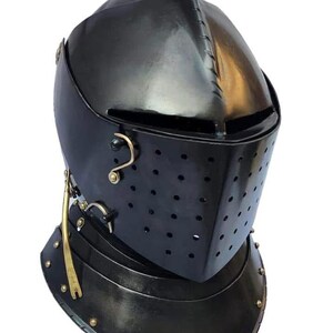 May include: A black metal knight helmet featuring a pointed visor with a horizontal eye slit and multiple ventilation holes. The helmet includes brass-coloured metal accents, including a side latch and small rivets along the neck guard base.