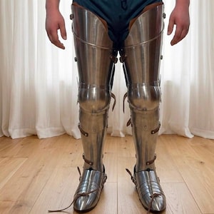 May include: A set of polished steel leg armor covering the thighs, knees, and shins, featuring articulated metal plates connected by brown leather straps and buckles. The armor extends down to include pointed metal sabatons for the feet.