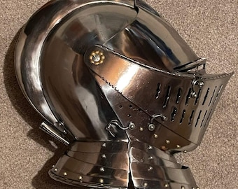 May include: A polished silver-toned metal close helmet featuring a rounded skull, a hinged visor with vertical ventilation slits, and a neck guard composed of overlapping lames secured with small rivets and a decorative brass-colored pivot point.
