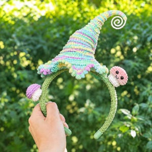 May include: A crochet headband featuring a tall, spiraled, pastel-colored pointed hat with a scalloped edge. Two small, crocheted mushroom figures with smiling faces are attached to the green, yarn-covered headband on either side of the hat.