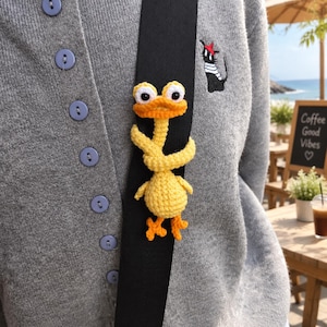 Può includere: Una figura di anatra all'uncinetto giallo brillante con grandi occhi bianchi e piedi arancioni, avvolta attorno a una tracolla nera. L'anatra è attaccata a un cardigan grigio abbottonato con un gatto ricamato in bianco e nero con un basco rosso.