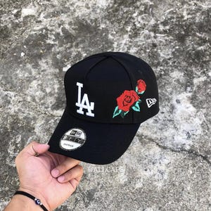 May include: Black baseball cap with the letters "LA" in white on the front. The cap features embroidered red roses with green leaves on the side. The New Era logo is visible. The cap is being held by a person.