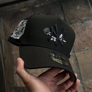 May include: Black baseball cap featuring the New York Yankees logo with a rose design. The cap has a curved brim and an adjustable strap. The side of the cap displays a patch with text. A stylish accessory for sports fans.