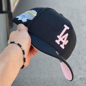 May include: Black baseball cap with a pink brim and the letters "LA" in pink. The cap features a patch with text and a multi-colored design. The cap is a fitted style.