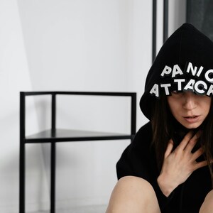 May include: Black hooded sweatshirt with the words "PANIC ATTACK" in white block letters across the hood. The person is wearing the hood and touching their neck. The background includes a black metal shelf and a white wall.
